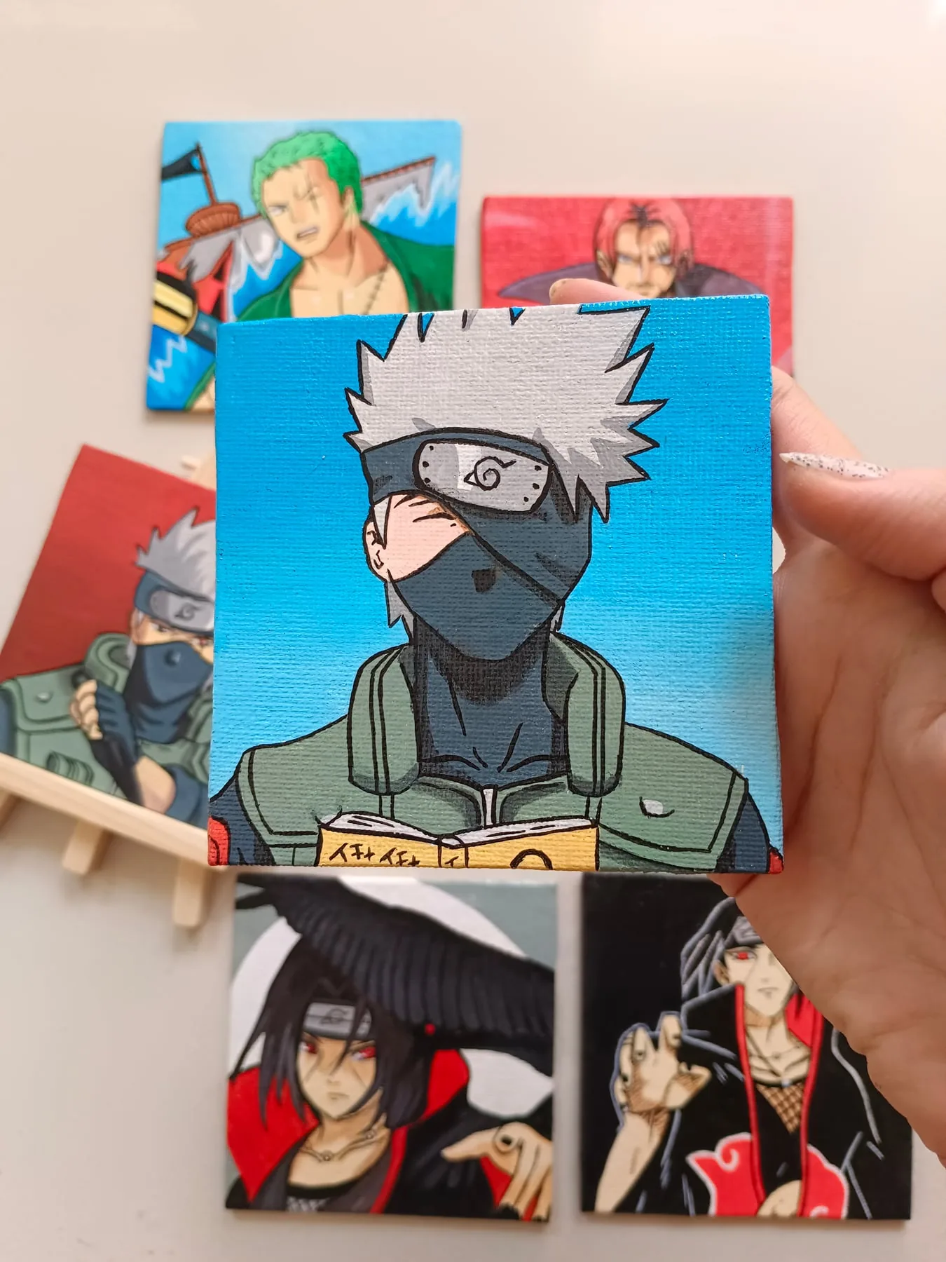 Hand-painted acrylic mini canvas of Kakashi Hatake from Naruto, held in front of other colourful character paintings.
