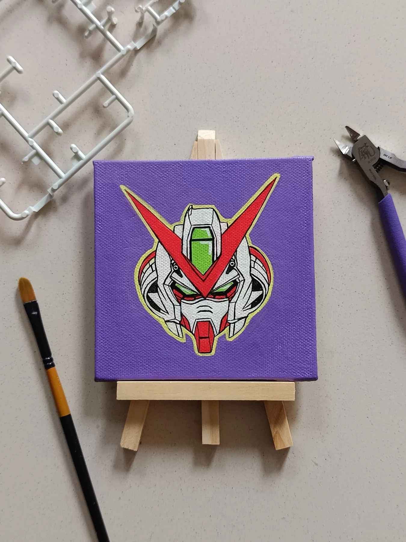 Hand-painted acrylic mini canvas of a Gundam-style mecha head on a purple background beside studio brushes.