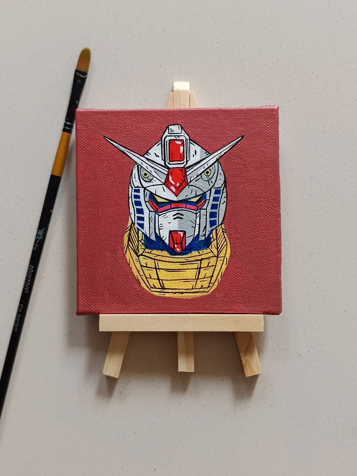 Hand-painted acrylic mini canvas of a Gundam-style mecha head on a red background, displayed on a small easel beside art brushes.