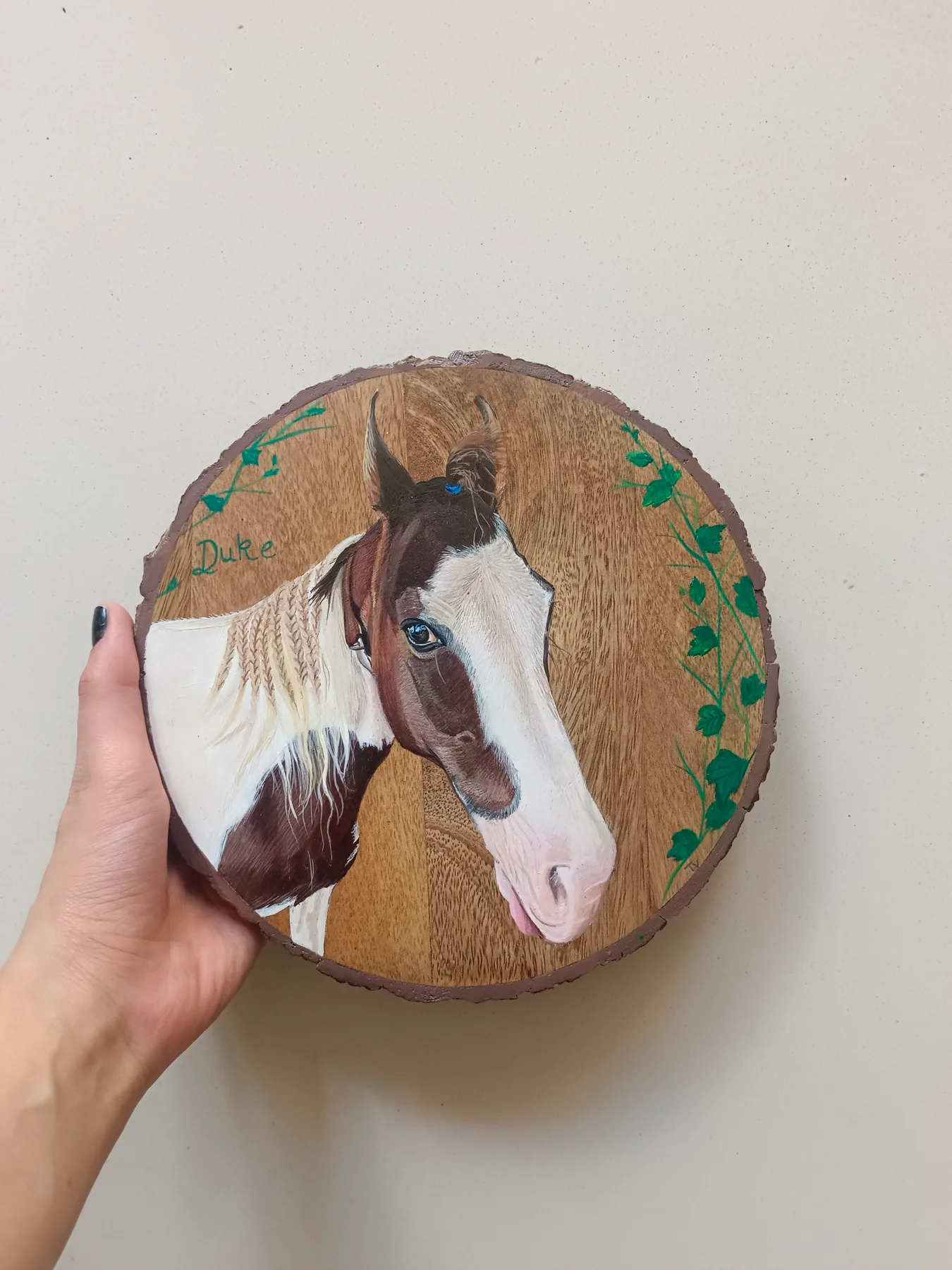 Round wood slice painting of a brown and white horse, held in hand with small green leaf details near the edge.