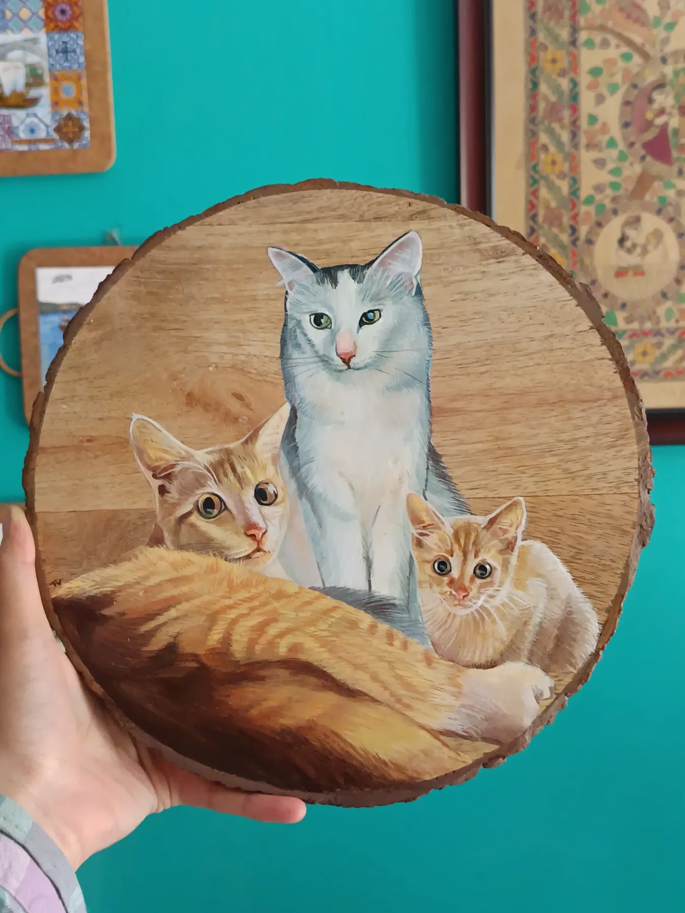 Round wood slice painting of three cats resting together, shown against a teal wall as a cosy pet portrait sample.