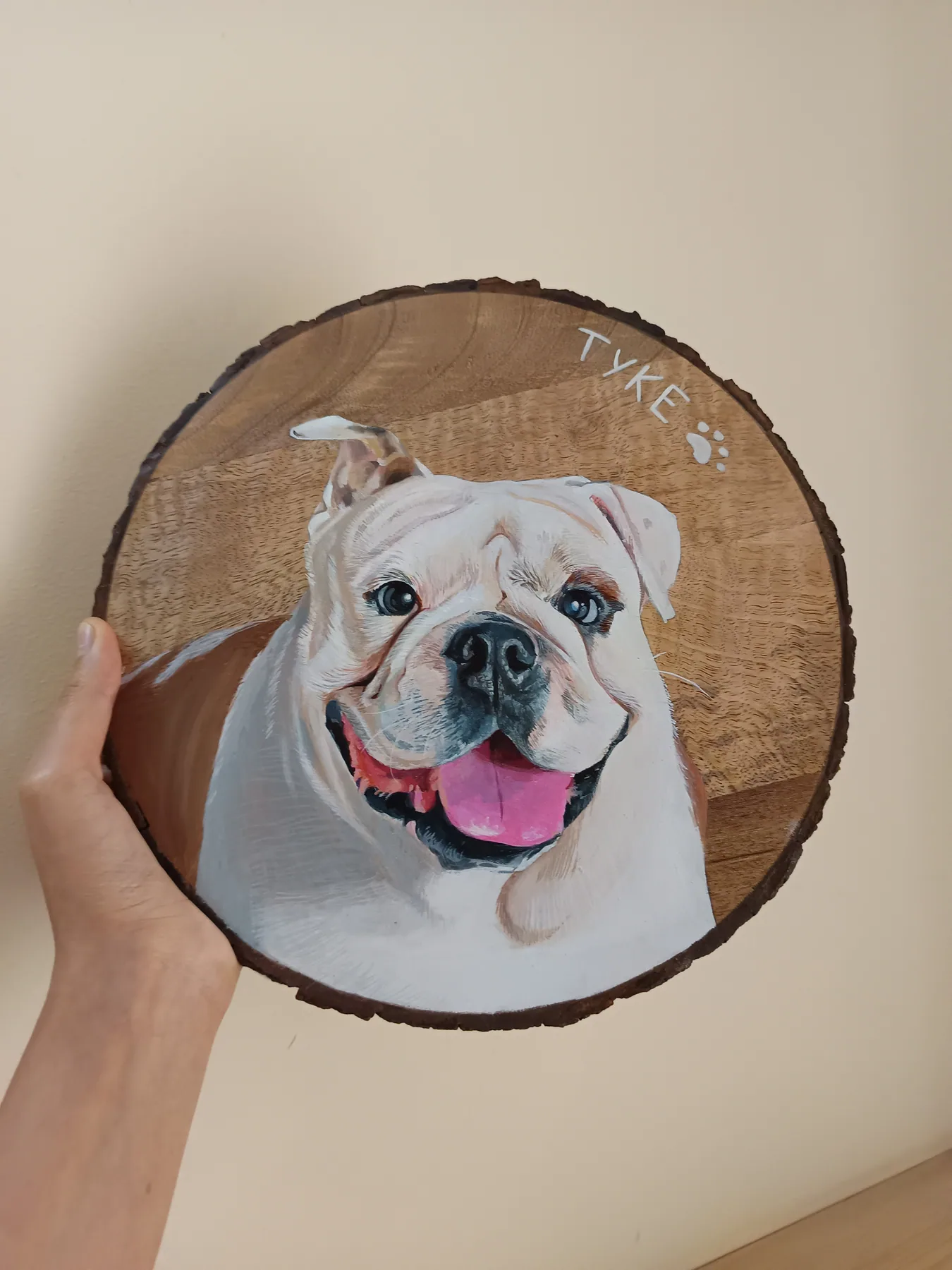 Round wood slice painting of a smiling white dog with a pink tongue, held in hand as a playful pet portrait sample.